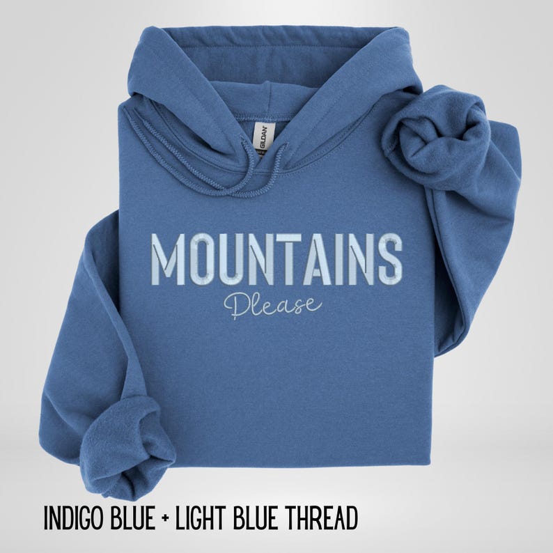 May include: A blue hooded sweatshirt with the words "MOUNTAINS Please" printed on the front in white. The sweatshirt is folded, with one sleeve rolled up. The text "INDIGO BLUE + LIGHT BLUE THREAD" is at the bottom.