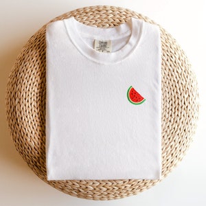 Puede incluir: Camiseta blanca con una peque&ntilde;a sand&iacute;a bordada en la parte delantera. La camiseta est&aacute; doblada y colocada sobre una estera de paja tejida.