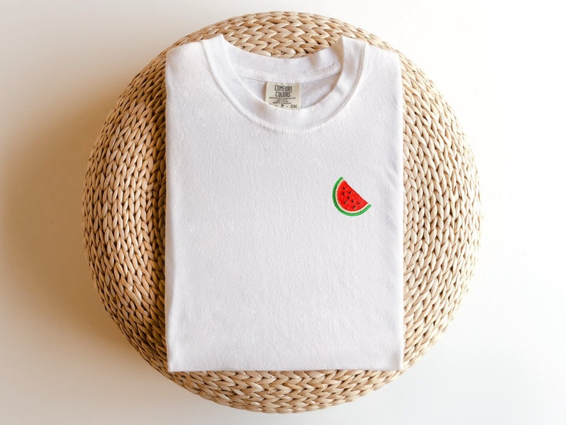 Puede incluir: Camiseta blanca con una peque&ntilde;a sand&iacute;a bordada en la parte delantera. La camiseta est&aacute; doblada y colocada sobre una estera de paja tejida.