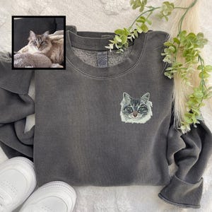 Puede incluir: Una sudadera gris oscuro con un rostro de gato bordado en blanco en la parte delantera. La sudadera est&aacute; hecha de un material suave y c&oacute;modo y tiene un corte holgado.
