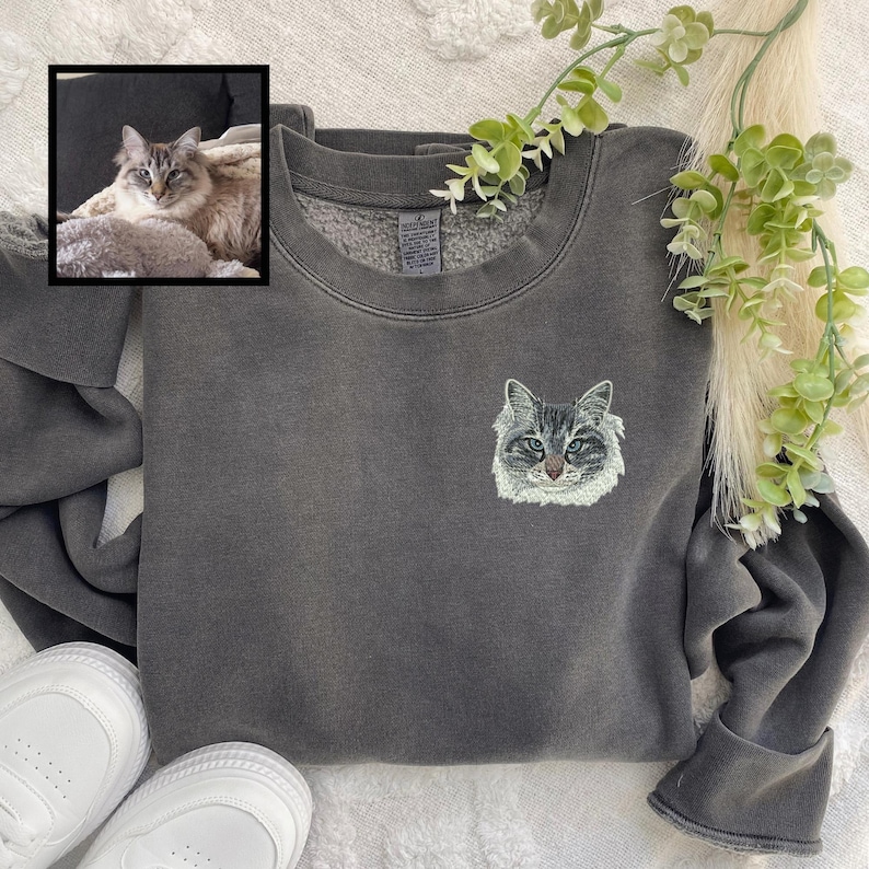 Puede incluir: Una sudadera gris oscuro con un rostro de gato bordado en blanco en la parte delantera. La sudadera est&aacute; hecha de un material suave y c&oacute;modo y tiene un corte holgado.