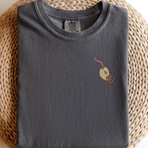 Puede incluir: Una camiseta gris oscuro con una peque&ntilde;a imagen bordada de una rata marr&oacute;n en la parte delantera. La camiseta tiene cuello redondo y mangas cortas.