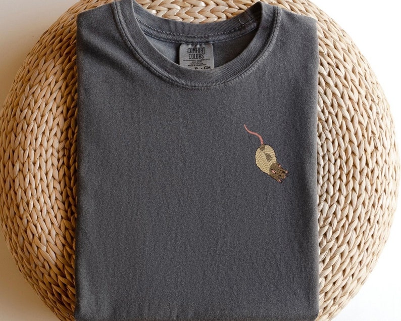 Puede incluir: Una camiseta gris oscuro con una peque&ntilde;a imagen bordada de una rata marr&oacute;n en la parte delantera. La camiseta tiene cuello redondo y mangas cortas.