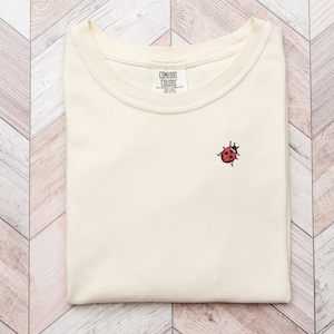 May include: A cream colored t-shirt with a small red ladybug embroidered on the front. The shirt has the text "COMFORT COLORS" on a tag.