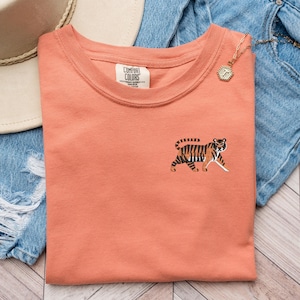 May include: A coral colored t-shirt with a small embroidered tiger on the left side of the chest. The t-shirt has a crew neck and is made of a soft, comfortable fabric. The brand name "Comfort Colors" is visible on the tag.