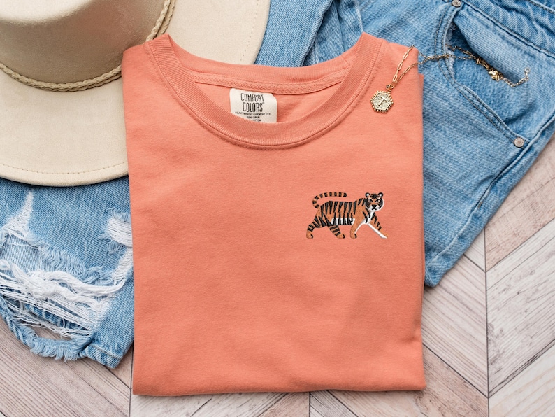 May include: A coral colored t-shirt with a small embroidered tiger on the left side of the chest. The t-shirt has a crew neck and is made of a soft, comfortable fabric. The brand name "Comfort Colors" is visible on the tag.
