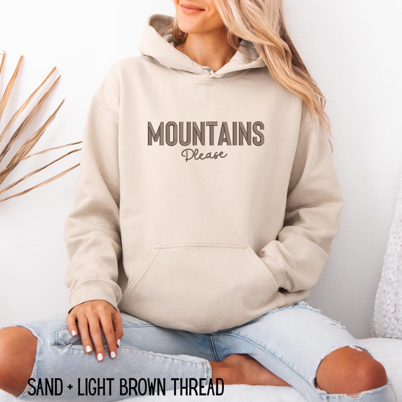 May include: A sand-colored hoodie with the text "MOUNTAINS Please" in light brown. The hoodie has a front pocket and a drawstring hood. The bottom of the image reads "SAND + LIGHT BROWN THREAD".