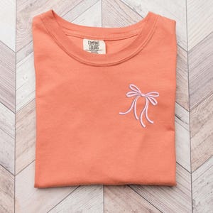 以下が含まれることがあります： 左側にピンクの刺繍リボンが付いたコーラルオレンジのTシャツ。Tシャツはクルーネックで、Comfort Colors製です。