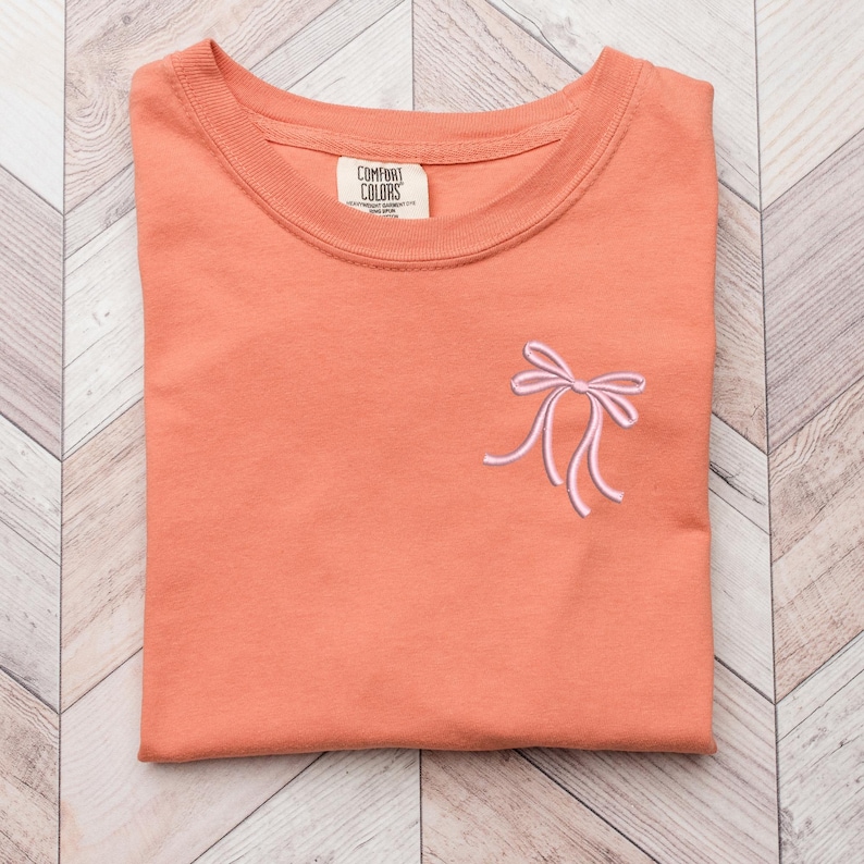 以下が含まれることがあります： 左側にピンクの刺繍リボンが付いたコーラルオレンジのTシャツ。Tシャツはクルーネックで、Comfort Colors製です。