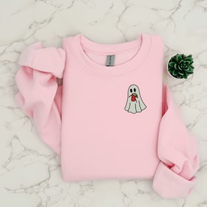 Puede incluir: Sudadera rosa claro de cuello redondo con un peque&ntilde;o dise&ntilde;o de fantasma bordado. El fantasma es blanco con un contorno negro y sostiene una fresa roja. La sudadera est&aacute; doblada sobre una superficie de m&aacute;rmol.