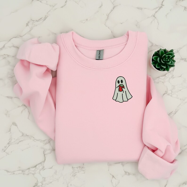 Puede incluir: Sudadera rosa claro de cuello redondo con un peque&ntilde;o dise&ntilde;o de fantasma bordado. El fantasma es blanco con un contorno negro y sostiene una fresa roja. La sudadera est&aacute; doblada sobre una superficie de m&aacute;rmol.