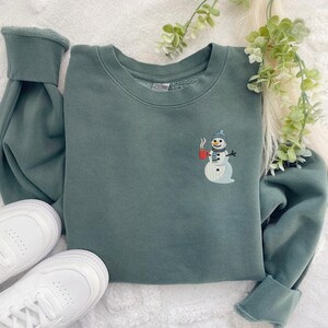 May include: A teal green sweatshirt with a crew neck and long sleeves. The sweatshirt features an embroidered snowman design on the front, holding a red mug. White sneakers are visible in the lower left corner.