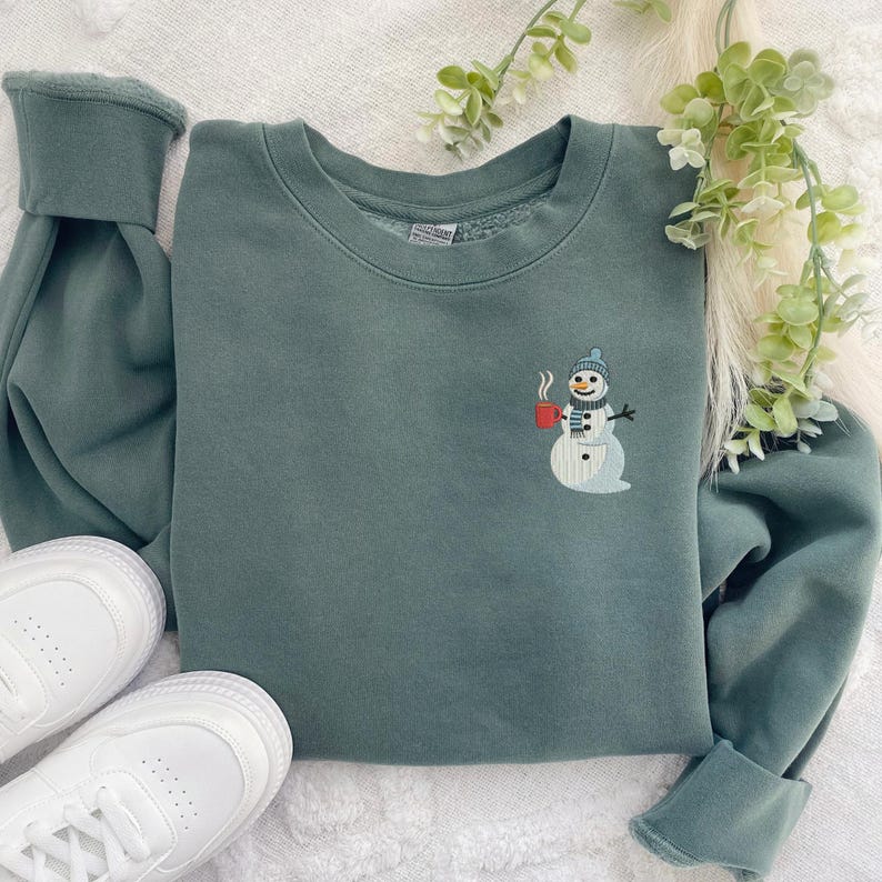May include: A teal green sweatshirt with a crew neck and long sleeves. The sweatshirt features an embroidered snowman design on the front, holding a red mug. White sneakers are visible in the lower left corner.