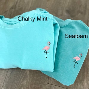 May include: Two pastel-colored sweatshirts, one in Chalky Mint and the other in Seafoam. Each sweatshirt features a small, embroidered pink flamingo on the front. The text "Chalky Mint" and "Seafoam" are visible on the sweatshirts.