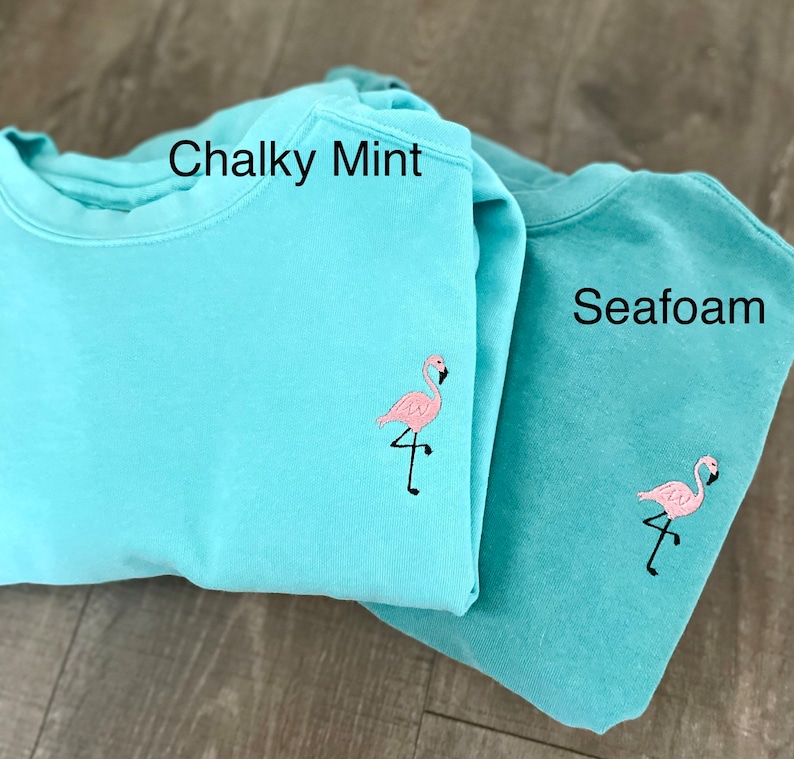 May include: Two pastel-colored sweatshirts, one in Chalky Mint and the other in Seafoam. Each sweatshirt features a small, embroidered pink flamingo on the front. The text "Chalky Mint" and "Seafoam" are visible on the sweatshirts.