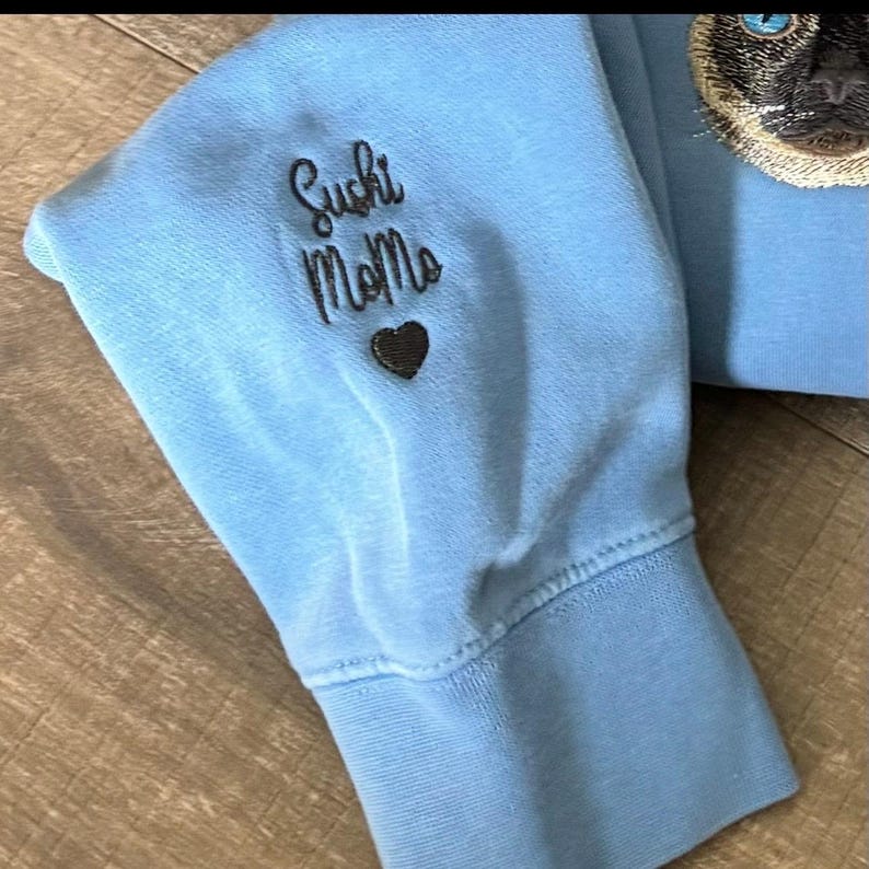 Puede incluir: Sudadera azul claro con un coraz&oacute;n bordado en negro y el texto "Sushi Momma" en la manga.