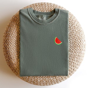 Puede incluir: Una camiseta verde con una peque&ntilde;a rodaja de sand&iacute;a bordada en la parte delantera. La camiseta est&aacute; doblada y colocada sobre una estera de paja tejida.