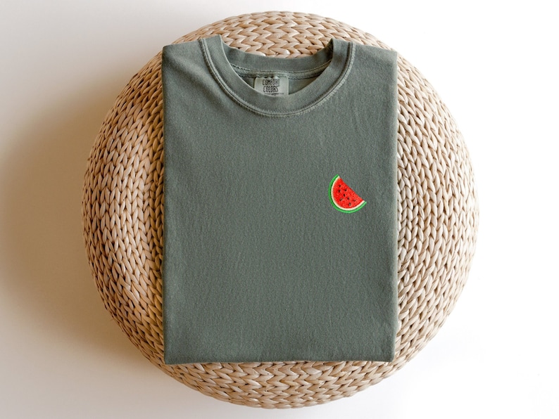 Puede incluir: Una camiseta verde con una peque&ntilde;a rodaja de sand&iacute;a bordada en la parte delantera. La camiseta est&aacute; doblada y colocada sobre una estera de paja tejida.