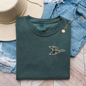 May include: A dark green t-shirt with a camouflage duck design. The shirt is laid out on a wooden surface with a pair of blue jeans and a brown hat.