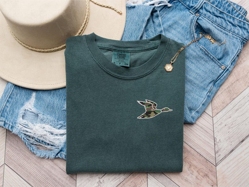 May include: A dark green t-shirt with a camouflage duck design. The shirt is laid out on a wooden surface with a pair of blue jeans and a brown hat.