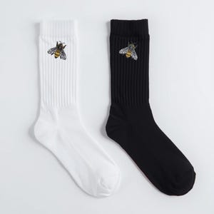 May include: A pair of crew socks, one white and one black, each featuring an embroidered bee design. The bee is detailed with yellow, black, and white accents. The socks are ribbed and appear to be made of a soft material.