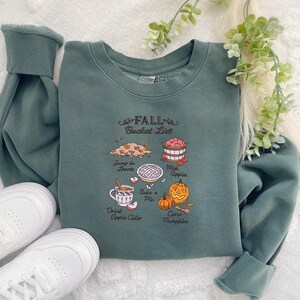 May include: A teal sweatshirt with the text "FALL Bucket List" and illustrations of fall activities. The illustrations include jumping in leaves, picking apples, baking a pie, drinking apple cider, and carving pumpkins. White sneakers are visible.