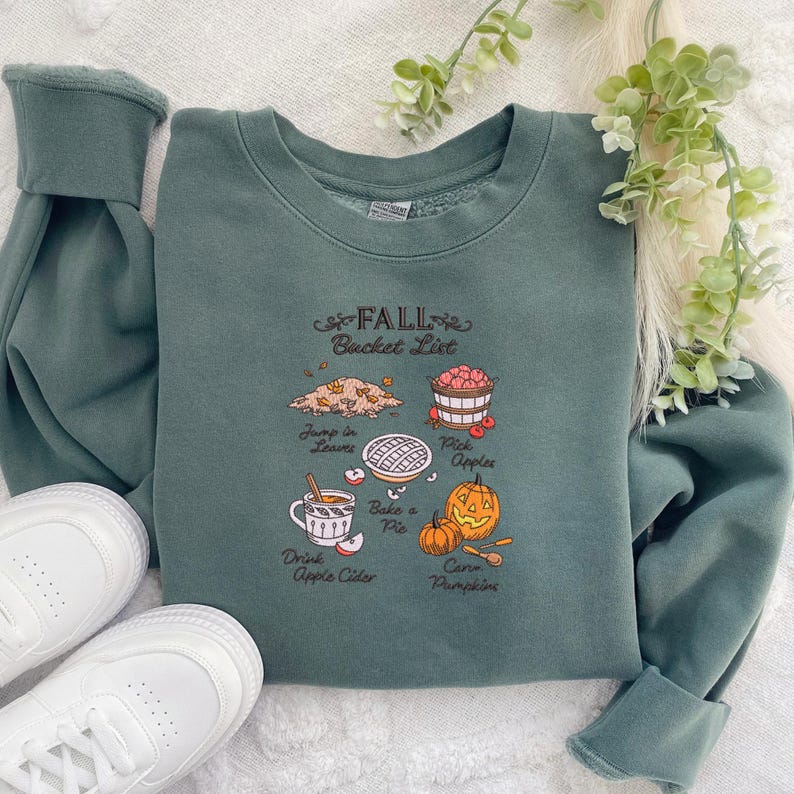 May include: A teal sweatshirt with the text "FALL Bucket List" and illustrations of fall activities. The illustrations include jumping in leaves, picking apples, baking a pie, drinking apple cider, and carving pumpkins. White sneakers are visible.