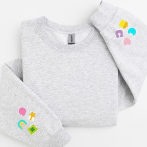 Peut inclure: Un sweat-shirt gris avec des patchs brod&eacute;s color&eacute;s sur les manches. Les patchs repr&eacute;sentent un arc-en-ciel, un tr&egrave;fle &agrave; quatre feuilles, une &eacute;toile, un c&oelig;ur, un ballon et un fer &agrave; cheval.