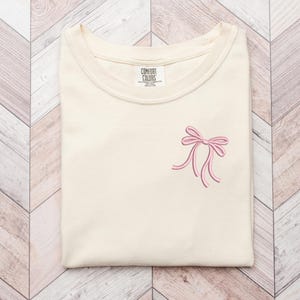 以下が含まれることがあります： クリーム色のTシャツに、ピンクの刺繍リボンが前面に施されています。Tシャツには「COMFORT COLORS」と書かれたタグが付いています。