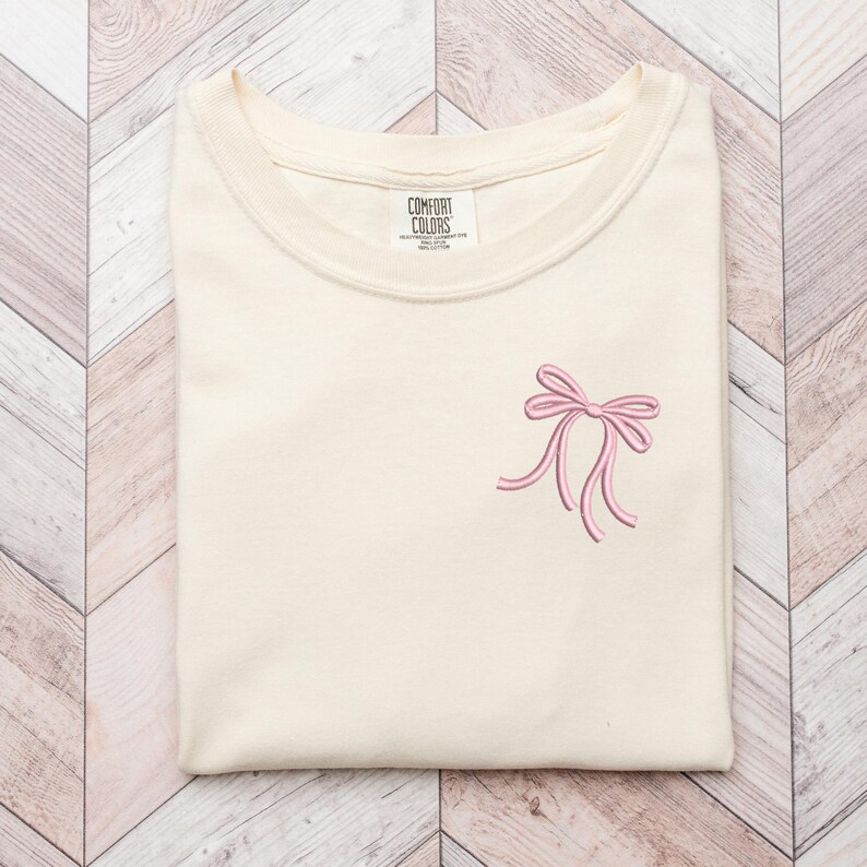 以下が含まれることがあります： クリーム色のTシャツに、ピンクの刺繍リボンが前面に施されています。Tシャツには「COMFORT COLORS」と書かれたタグが付いています。
