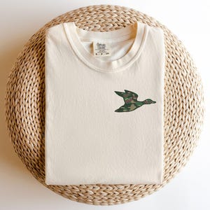 May include: A cream colored t-shirt with a camouflage duck design on the front. The t-shirt is on a woven straw mat.