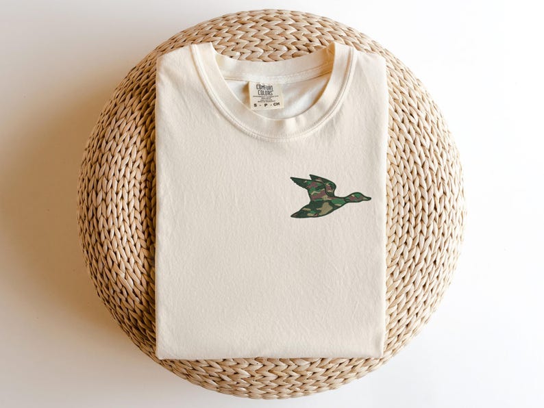 May include: A cream colored t-shirt with a camouflage duck design on the front. The t-shirt is on a woven straw mat.