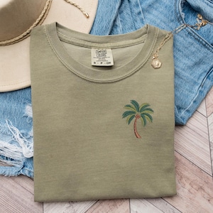 Op de afbeelding: Een groen T-shirt met een geborduurde palmboom op de voorkant. Het T-shirt heeft een ronde hals en is gemaakt van een zachte, comfortabele stof. Het label op het shirt luidt "Comfort Colors".
