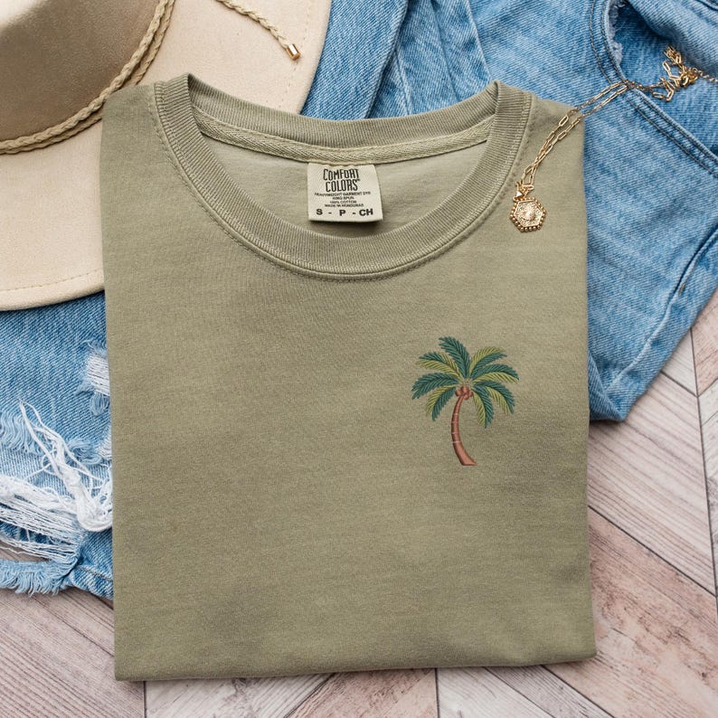 Op de afbeelding: Een groen T-shirt met een geborduurde palmboom op de voorkant. Het T-shirt heeft een ronde hals en is gemaakt van een zachte, comfortabele stof. Het label op het shirt luidt "Comfort Colors".