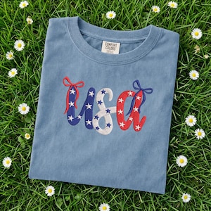 Pu&ograve; includere: Una maglietta blu Comfort Colors con ricami "USA". Le lettere sono decorate con stelle e fiocchi rossi, bianchi e blu, evocando un tema patriottico. La maglietta &egrave; adagiata su un letto di erba verde con piccoli fiori bianchi.