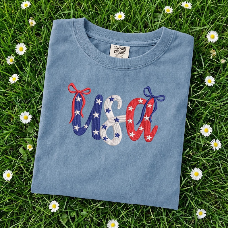 Pu&ograve; includere: Una maglietta blu Comfort Colors con ricami "USA". Le lettere sono decorate con stelle e fiocchi rossi, bianchi e blu, evocando un tema patriottico. La maglietta &egrave; adagiata su un letto di erba verde con piccoli fiori bianchi.