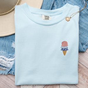 May include: Light blue t-shirt with an embroidered ice cream cone design. The ice cream features red and white stripes, blue stars, and a brown cone. The shirt has a Comfort Colors tag.