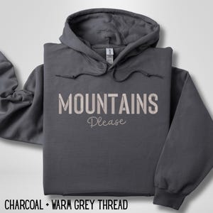 May include: A charcoal gray hoodie with the words "MOUNTAINS Please" in a warm gray thread. The hoodie has a drawstring hood and a front pocket. The text "CHARCOAL + WARM GREY THREAD" is at the bottom.