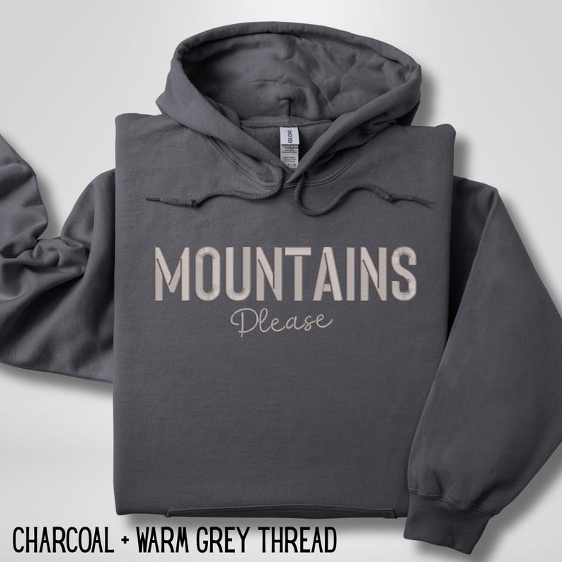 May include: A charcoal gray hoodie with the words "MOUNTAINS Please" in a warm gray thread. The hoodie has a drawstring hood and a front pocket. The text "CHARCOAL + WARM GREY THREAD" is at the bottom.