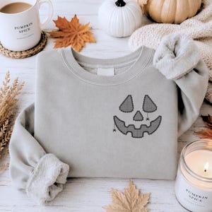 Puede incluir: Sudadera gris claro con un diseño de cara de calabaza negra en la parte delantera. El diseño incluye ojos triangulares, una nariz y una boca sonriente con acentos de araña. La sudadera tiene mangas largas y cuello redondo.