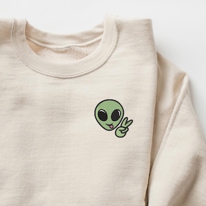 May include: A beige sweatshirt with a green alien cartoon graphic wearing a peace sign. The alien is sticking its tongue out.