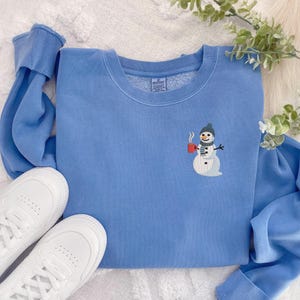 May include: A light blue sweatshirt with a crew neck and long sleeves. The sweatshirt features an embroidered snowman design on the front, holding a red mug. White sneakers are visible in the bottom left corner.
