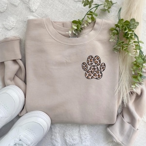 May include: A beige sweatshirt with a leopard print paw print design. The sweatshirt is made of a soft, comfortable material and is perfect for everyday wear.