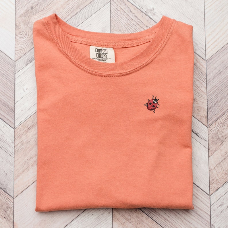 Puede incluir: Una camiseta de color coral doblada con una peque&ntilde;a mariquita bordada en la parte delantera. La camiseta tiene una etiqueta que dice "COMFORT COLORS" y "HEAVYWEIGHT GARMENT DYE".
