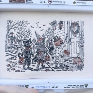 May include: An embroidery design on off-white fabric featuring three figures in Halloween costumes: a black cat, a witch, and a pumpkin-headed character ringing a doorbell. The scene includes a crescent moon, bare trees, a house, and pumpkins, all rendered in black and orange thread within an 8x13 inch embroidery hoop.