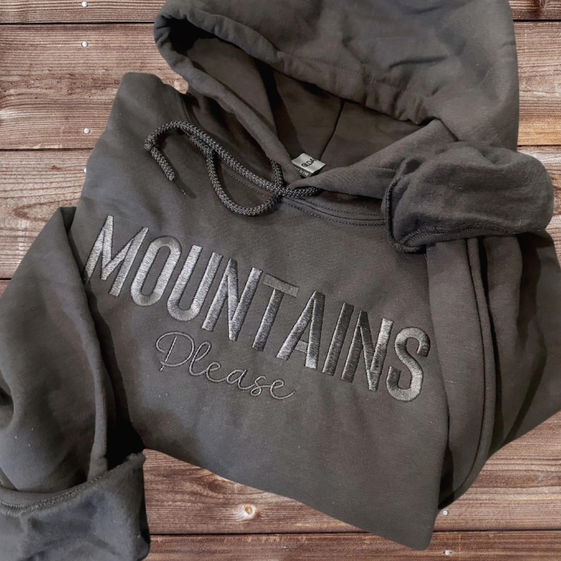 May include: Dark gray hoodie with the embroidered text "MOUNTAINS Please". The hoodie has a drawstring hood and a front pocket. The garment is made of a soft material and is folded on a wooden surface.