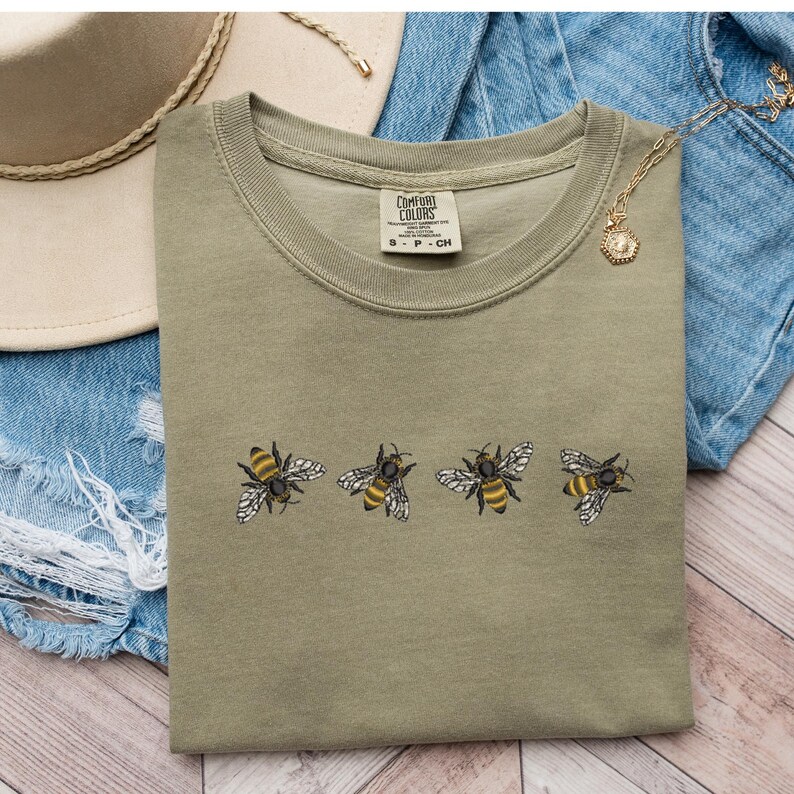 Może przedstawiać: Zielony T-shirt z rzędem czterech haftowanych pszcz&oacute;ł. Pszczoły są ż&oacute;łte i czarne z brązowymi skrzydłami. Koszulka ma metkę z napisem "Comfort Colors" i "S - P - CH".