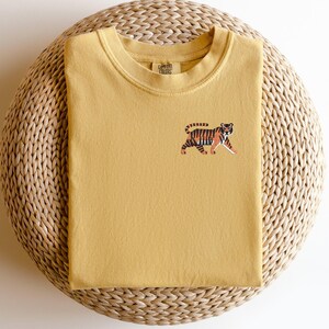 May include: A mustard yellow t-shirt with an embroidered tiger design. The t-shirt has a crew neck and is made of a soft, comfortable fabric.