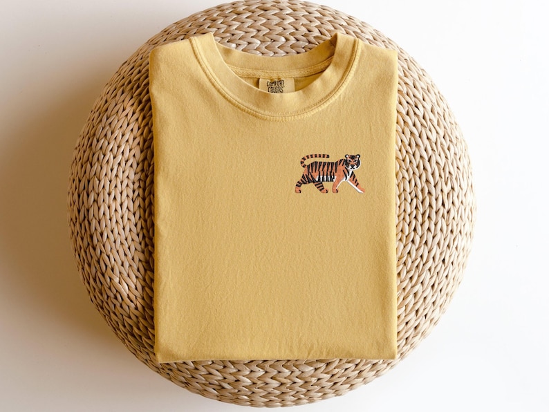 May include: A mustard yellow t-shirt with an embroidered tiger design. The t-shirt has a crew neck and is made of a soft, comfortable fabric.