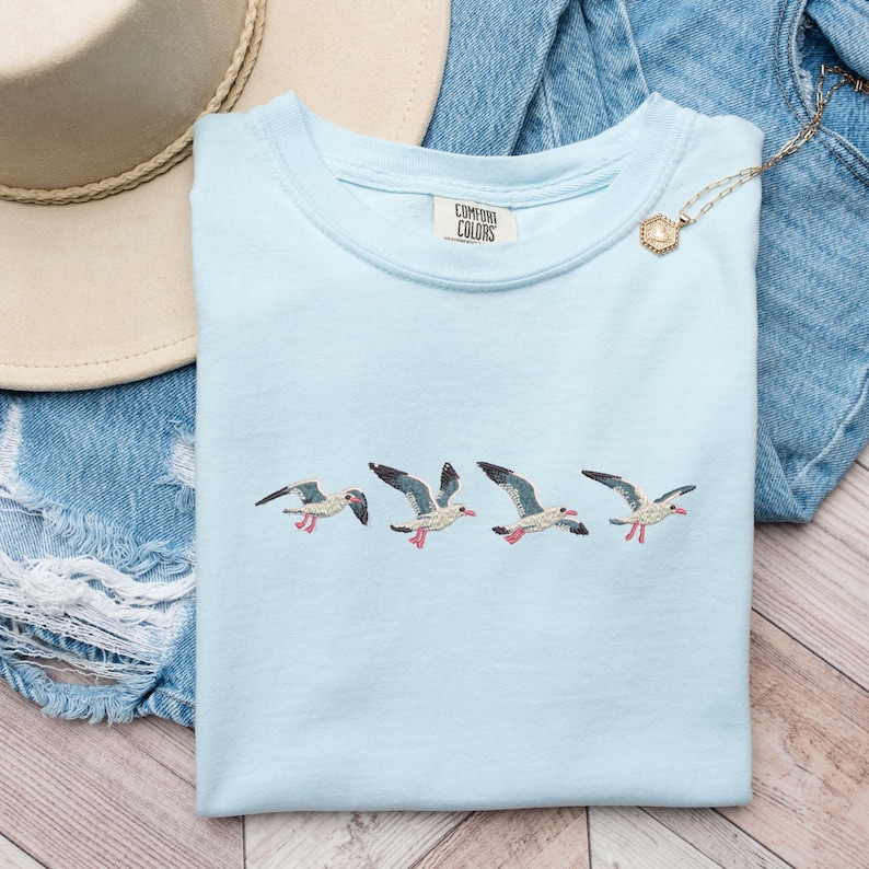 Puede incluir: Camiseta azul claro con gaviotas bordadas. La camiseta tiene el texto "Comfort Colors" en la etiqueta.